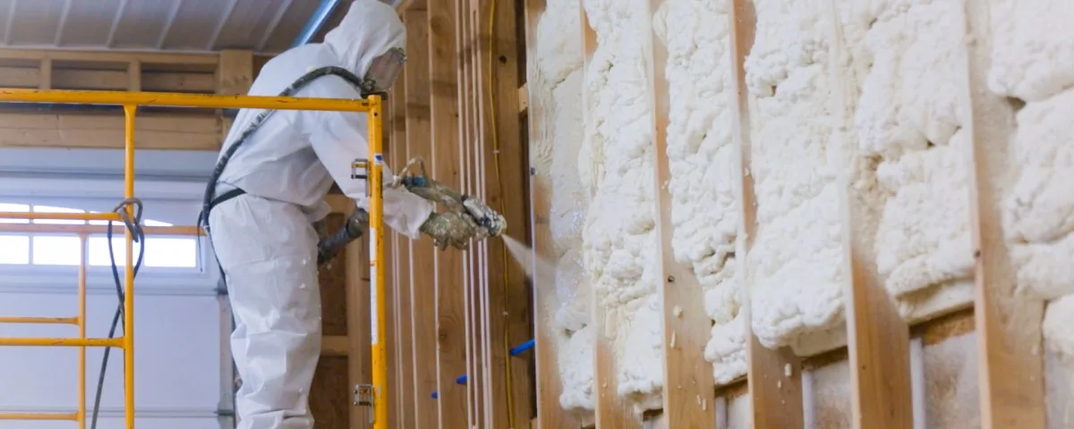 Spray foam operator in full PPE — Tyvek coverall, supplied-air respirator hood, butyl gloves — spraying PU foam onto a wall